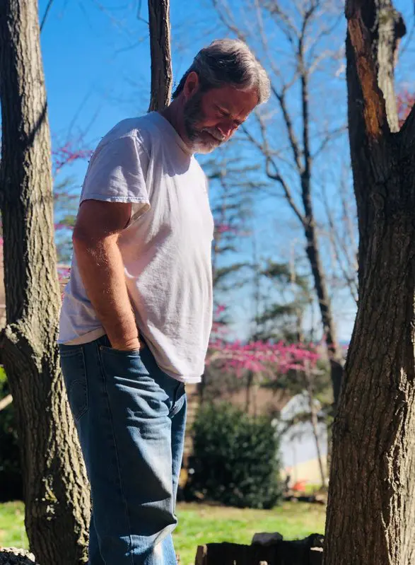 A man in a white t-shirt and jeans stands outdoors between trees.